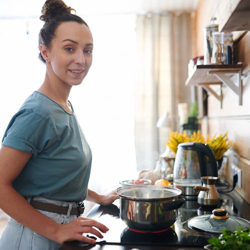 Young pretty housewife in t-shirt and jeans looking at you while standing by electric stove in the kitchen