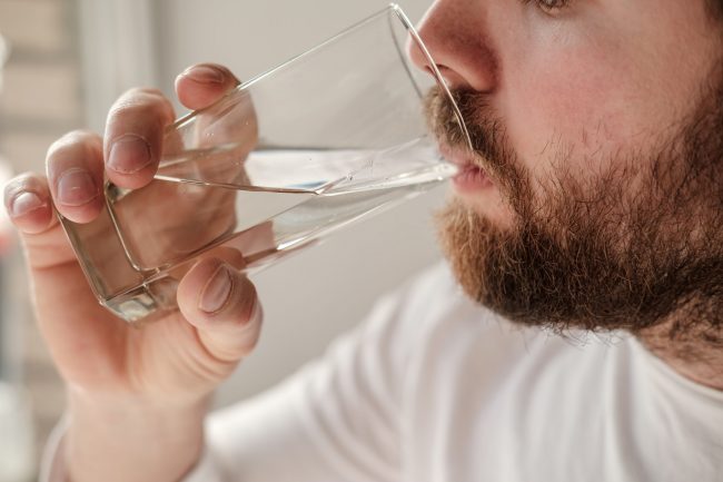 Close-up of a handsome bearded man drinking clear water from a transparent glass. Health concept.